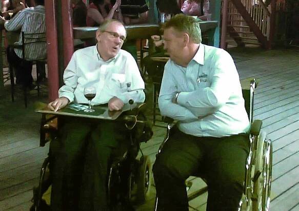 Two male wheelchair users having a chat in a pub. One has a tray and glass of wine.