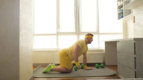 Domácí cvičení na fitness podložce, pumpovat záda s činkami doma ve zpomaleném filmu. Mladý sportovec napumpuje svaly na pažích v bytě. — Stock záběr