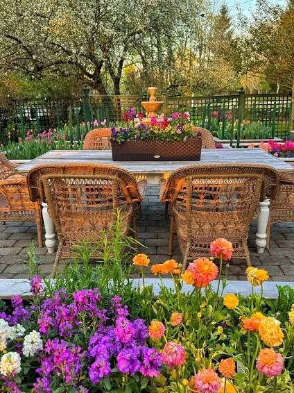 Wicker chairs surround a white table on a stone patio, with colorful flowers in the foreground and a planter centerpiece. Lush green trees and blooming flowers fill the background, evoking a serene garden setting.