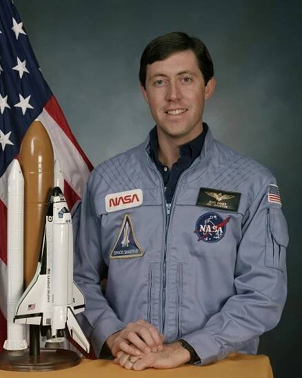 A man stands in front of the American flag and next to him is a small rocket figurine 