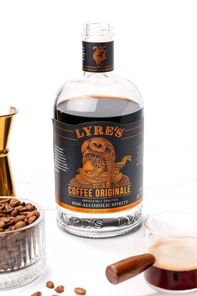 A bottle of Lyre's Coffee Originale non-alcoholic spirits is placed on a surface alongside a metal jigger, a bowl of coffee beans, and a cup of espresso.