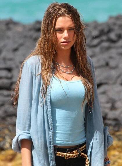a woman with long hair standing next to the ocean wearing a blue shirt and black pants