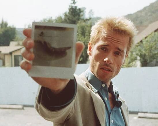 A man with blonde spiky hair holds up a Polaroid photograph.