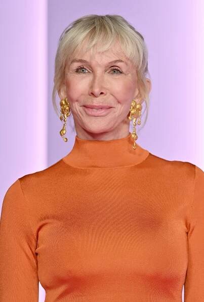 At the Green Carpet Fashion Awards in Hollywood, Trudie Styler stuns in a bold orange high-neck gown paired with sculptural gold earrings, showcasing vibrant style, confidence, and modern elegance.