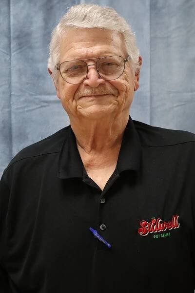 An older man wearing glasses and a black shirt is standing in front of a blue curtain.