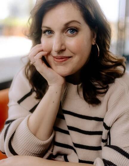 A woman in a striped sweater smiles at the camera with her chin in her hand