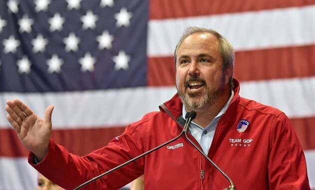 Joe Gruters speaking at a podium in front of an American flag.