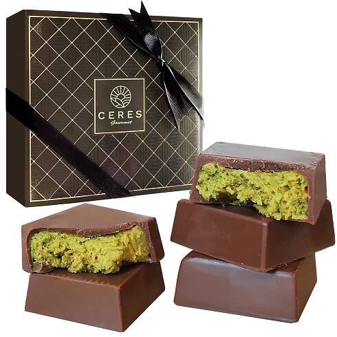 A Ceres Gourmet gift box with a black ribbon sits behind three Dubai Knafah Pistachio Chocolate Bar bites, each filled with green pistachio kunafa; one is split to reveal the inside. The box contains 16 exquisite chocolate bite sizes.