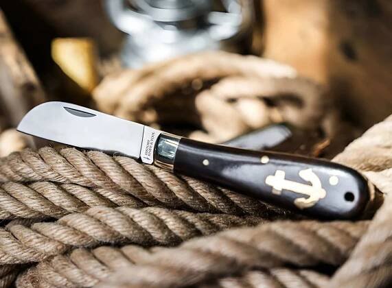 A folding knife with a polished metal blade and a dark wooden handle featuring a gold anchor inlay, resting on coiled thick rope. The blade is engraved with the text "HISTOIRE KNIFE & TOOL.