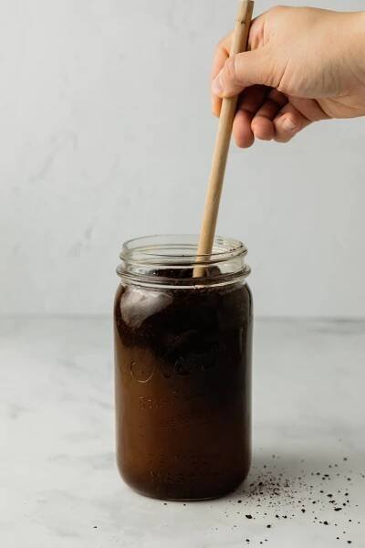 A hand holds a wooden spoon and stirs a mixture of coffee grounds and water.