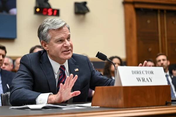 Christopher A. Wray, wearing a dark suit and striped tie, sitting at a table with a microphone in front of him. 