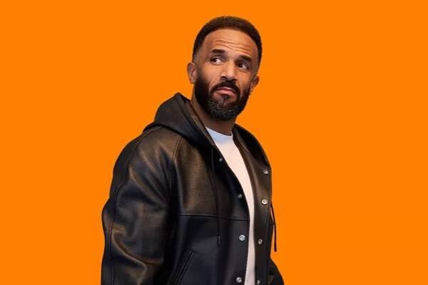 Craig David, in a white t-shirt and black jacket, with an orange background behind him in a promotional photo for Just Eat.