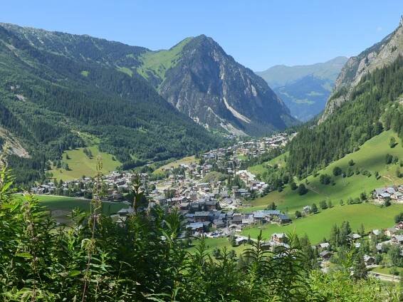 Blick auf ein malerisches Tal mit einer kleinen Stadt inmitten von grünen Hügeln und hohen Bergen unter klarem, blauem Himmel.
