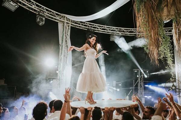 Bride Inbar Lavi smiling and posing as guests lift her up on a table at the wedding reception