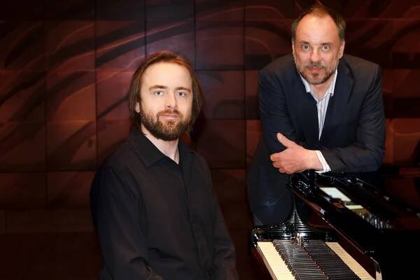 Portrait von Daniil Trifonov und Matthias Goerne. Mann mit längeren Haaren, Vollbart und shcwarzer Kleidung sitzt an einem Klavier. Mann mit schütterem Haar und grauem Bart steht daneben und lehnt am Klavier. Beide blicken lächelnd in die Kamera