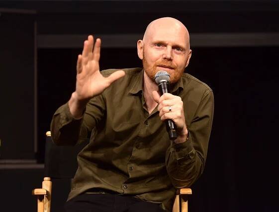 An image of Bill Burr wearing a brown shirt, holding one hand up, and speaking into a microphone for our article about whether he'll reprise his Breaking Bad role on Better Call Saul