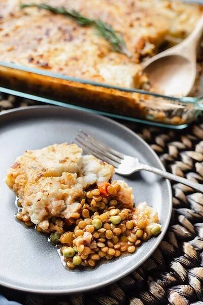 vegan lentil shepherd's pie overlay in a rectangular glass serving dish with a spoon with a serving in a grey plate on a brown background