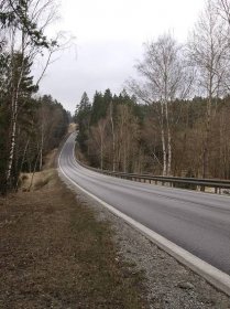 Silnice I/3 v místě nehody, pohled z levé krajnice směrem k můstku přes Hněvanovický potok. Autobus přijížděl z protějšího kopce ze směru od Dolního Dvořiště (duben 2018)