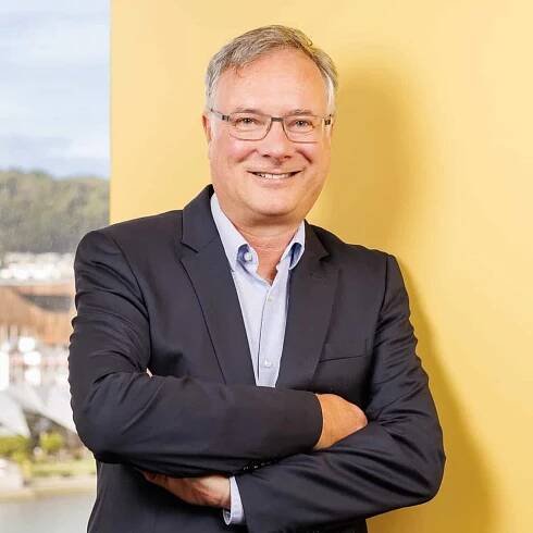 A middle-aged man with gray hair and glasses is smiling at the camera. He is wearing a dark blazer over a light blue shirt and has his arms crossed. The background is yellow with a window on the left showing an outdoor scene.