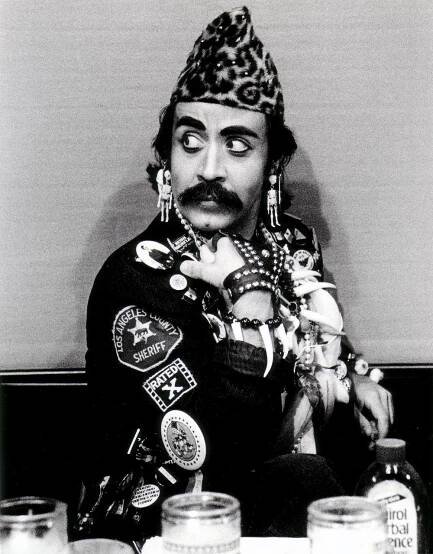 Guillermo Gómez-Peña sitting at an altar decorated with a kitsch collection of cultural fetish items, and wearing a border patrolman’s jacket decorated with buttons, bananas, beads, and shells.