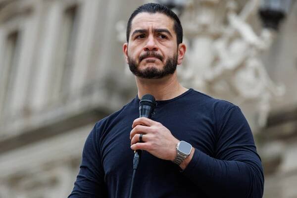 Abdul El-Sayed stands outside in front an imposing government building. He holds a black microphone and appears to be scowling.