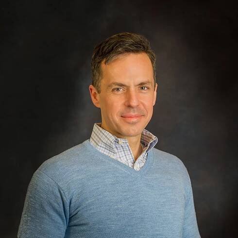 Chris Moore stands in front of a dark grey background. He wears a long-sleeve blue top with a white checkered button-up underneath.