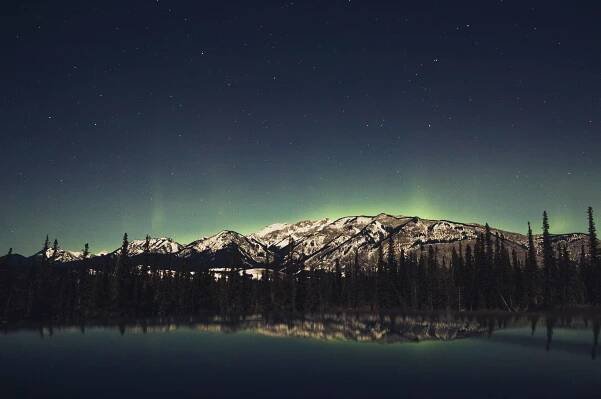 the green northern lights dance behind a mountain againast a starry night sky
