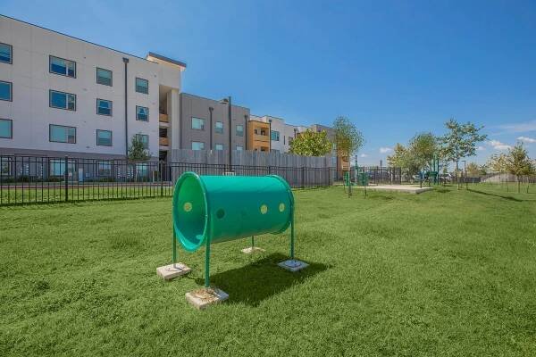A brightly colored green play tunnel sits on a grassy area in a park. In the background, there are modern multi-story residential buildings. The sky is clear and blue, and there are a few trees scattered around the space, adding to the inviting atmosphere of the park.