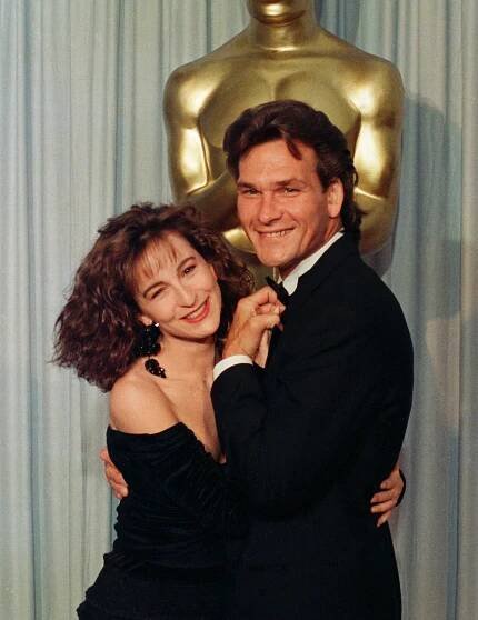 Jennifer Grey and Patrick Swayze smiling, standing together in front of an Oscar statue. The woman wears a off-the-shoulder dress, and the man wears a tuxedo