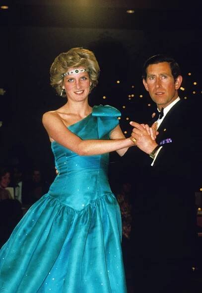 Princess Diana wearing a green emerald headband and aquamarine ballgown, holding hands with Prince Charles who is wearing a tuxedo.