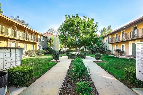 A well-maintained courtyard in an apartment community featuring green grass, neatly trimmed shrubs, and a central tree. Surrounding the pathway are two-story buildings with balconies, and there are outdoor mailboxes visible on one side. The scene is bright and inviting, with clear blue skies.