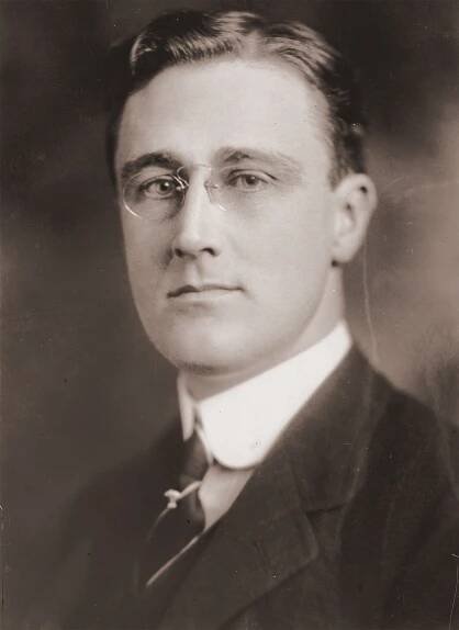 Vintage sepia-toned portrait of a young Franklin D. Roosevelt, wearing round eyeglasses and a suit with a white collared shirt and tie. Roosevelt is depicted as Assistant Secretary of the Navy, taken between 1909 and 1919.