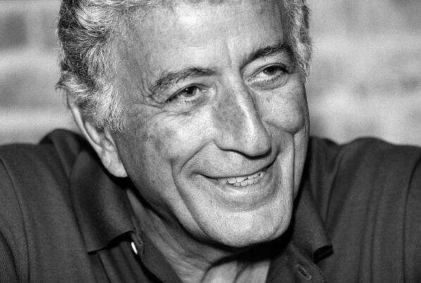 A close-up black and white photo of Tony Bennett in his late 60s, wearing a dark open-collared shirt and a gentle smile. 