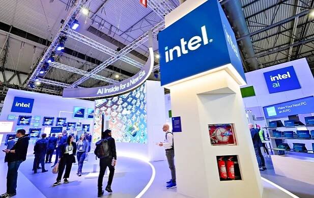 Trade show booth with Intel branding and digital displays. Visitors explore AI technology exhibits. Large Intel signs are prominent, and fire extinguishers are visible on a white pillar. The atmosphere is modern and tech-focused.