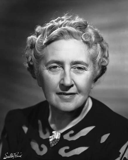Elderly woman with curled hair wearing a dark top and a decorative brooch, smiling softly in a portrait-style photograph