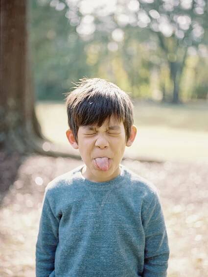 Young child in blue sweater making silly face with tongue out in outdoor setting.