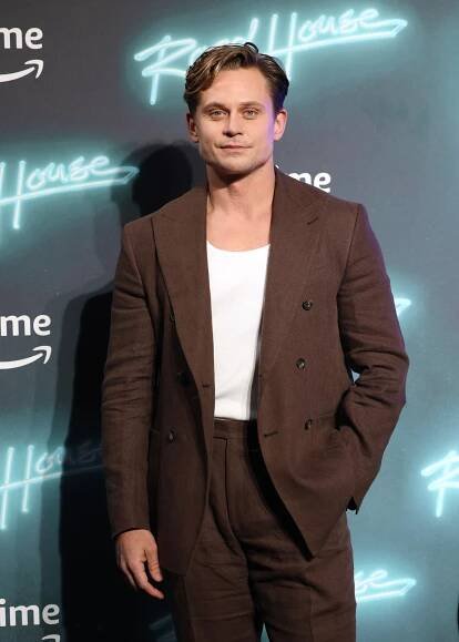 Billy Magnussen at the "Road House" event, wearing a double-breasted brown suit with a white shirt underneath, standing in front of the event backdrop