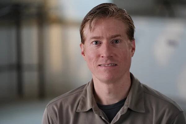 Portrait of a person with short, light hair, wearing a beige collared shirt over a gray top, in front of a blurred indoor background.
