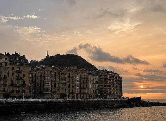 A sunset over a waterfront cityscape in San Sebastian, Spain, with historic buildings lining the shore. In the background, a statue stands atop a hill under a cloudy sky, and the sun sets on the horizon, casting a warm glow over this must-see destination among places to visit in Spain in 2025.