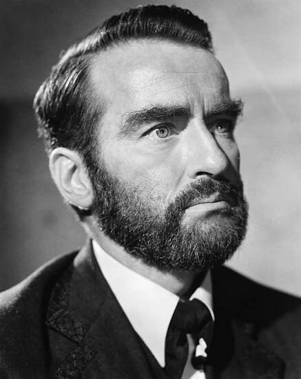 A person with a beard and styled hair in a formal suit looks contemplatively into the distance