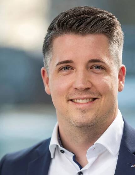 Ein Mann mit kurzen braunen Haaren und blauen Augen, der eine marineblaue Anzugsjacke und ein weißes Hemd trägt, lächelt in die Kamera. Der unscharfe Hintergrund im Freien deutet auf ein professionelles Umfeld hin, perfekt für die Präsentation von Karrieren oder Jobs bei Repabad.