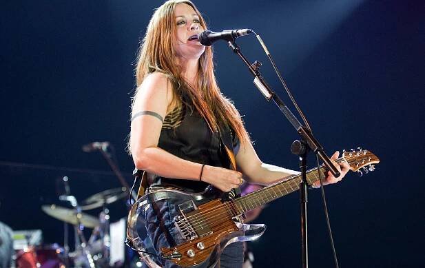A woman in jeans and a black vest, holding a transparent guitar and standing behind a microphone stand. In the background is a drum set.