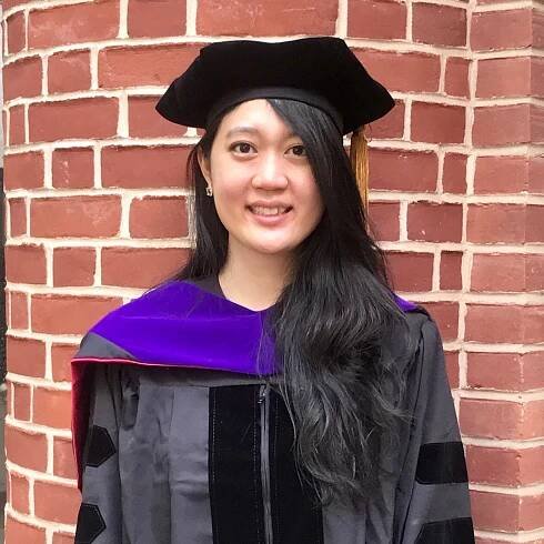 Sarah Lee Best standing in front of a brick wall and wearing a dark graduation cap and gown 