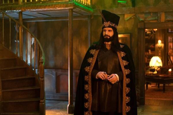 Kayvan Novak stands in a dark room wearing a hat and cloak as the vampire Nandor in "What We Do in the Shadows" Season 3.