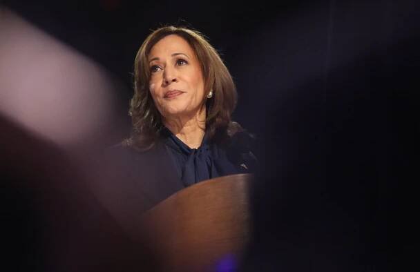 Kamala Harris standing at a podium in a spotlight wearing a blue blazer and blue blouse and looking up and to her right.