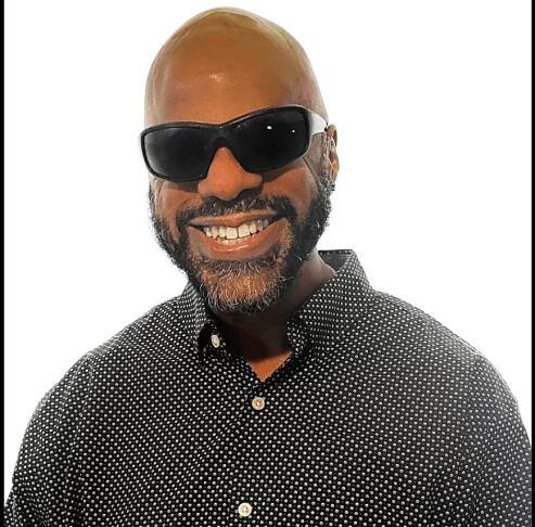 Graphic with a black background featuring a photo of Thomas Reid, a brown skin Black man with a clean-shaven bald head and full neat beard,  smiles into the camera. He wears dark shades and a black and white checkered button up shirt.