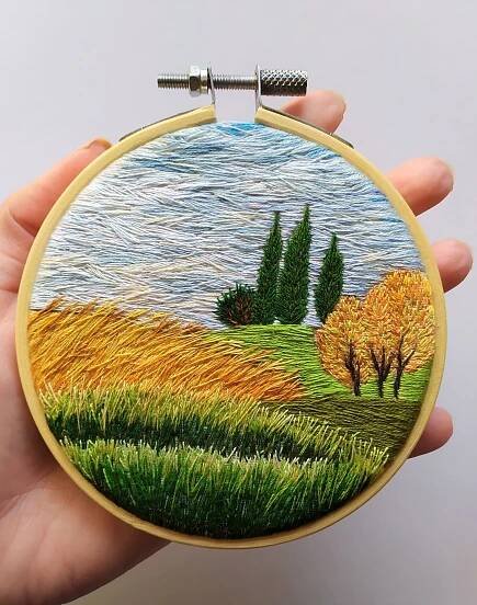 a hand holding up a small embroidery art piece in front of a white background with trees and grass