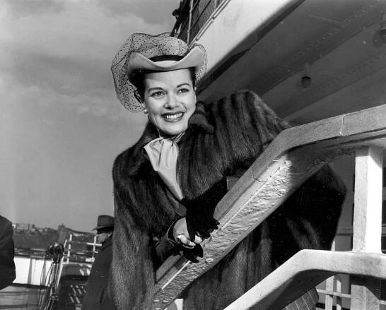 A black and white photo of Janis Paige wearing a hat and fur coat and leaning over a railing while smiling.