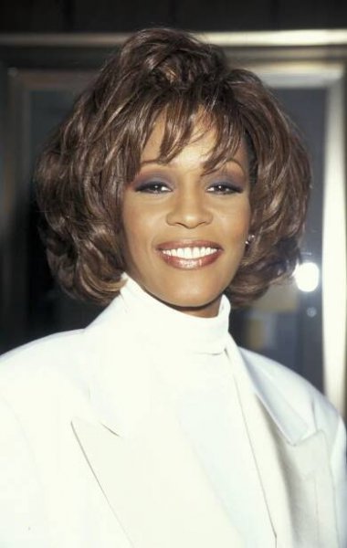 Person smiling in elegant attire with a high-neck top and blazer, looking at the camera against an indoor background