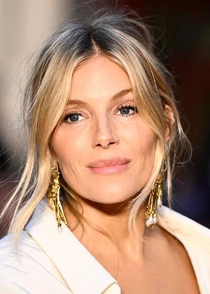 Sienna Miller wears subtle face-framing layers, dangly gold earrings, and a cream shirt